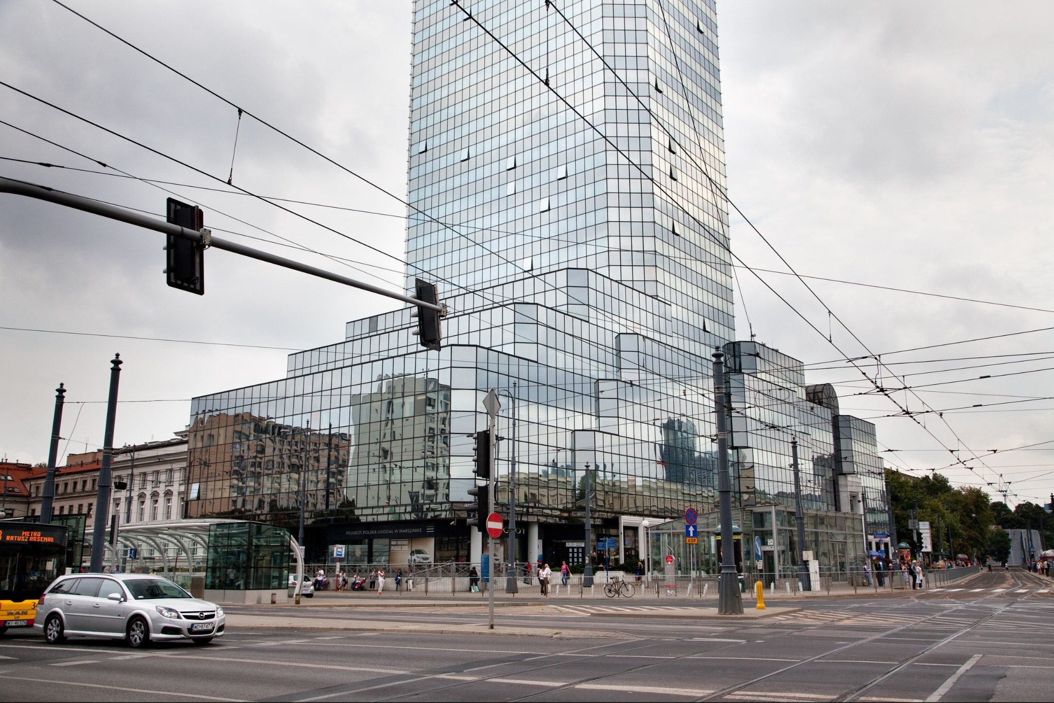 Szklany biurowiec w miejskiej scenerii z ulicą, tramwajami i przechodniami w pochmurny dzień.