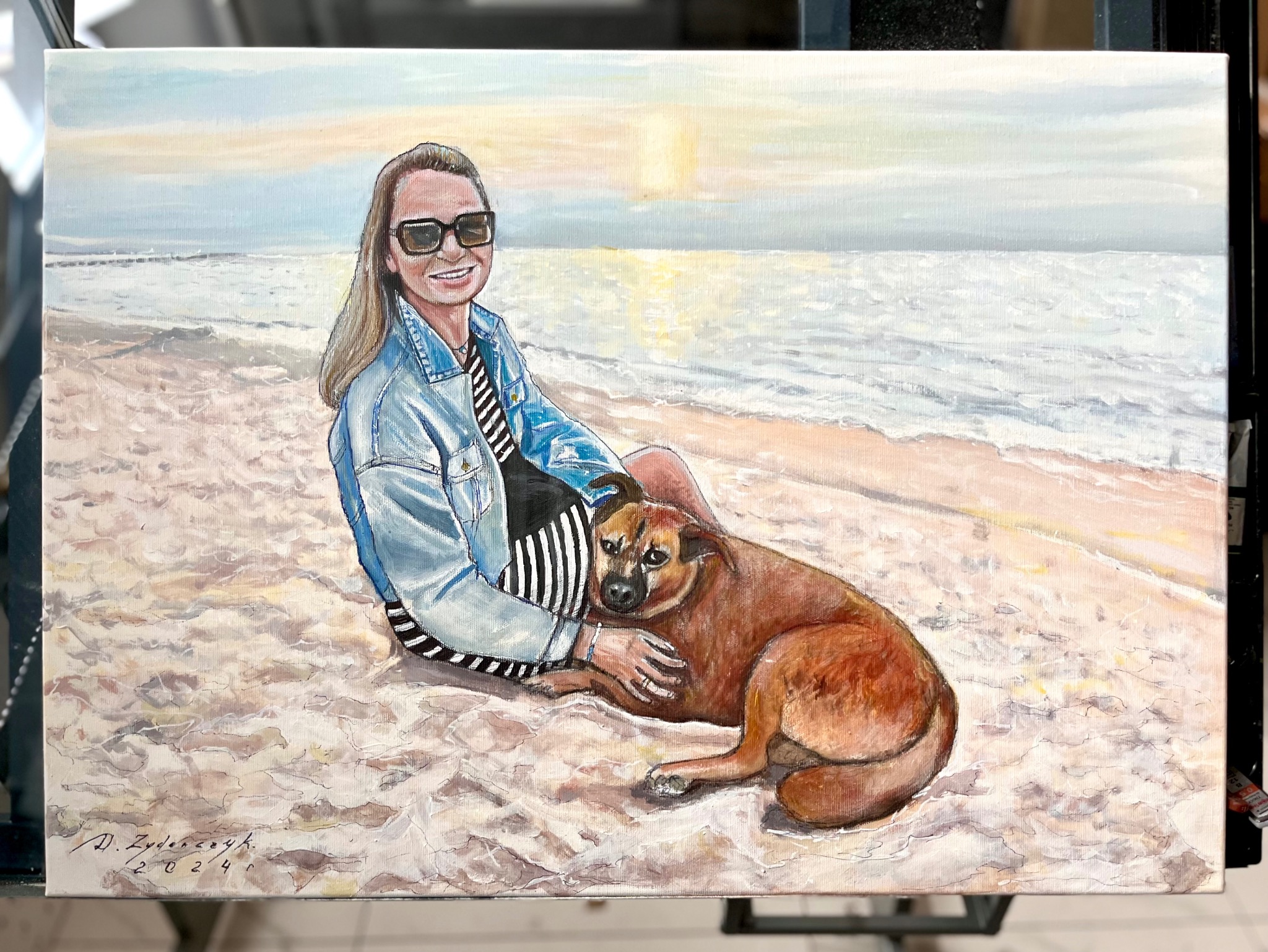 Obraz olejny przedstawiający uśmiechniętą kobietę w okularach przeciwsłonecznych i dżinsowej kurtce, siedzącą na plaży z psem, z podpisem autora i datą 2024 w lewym dolnym rogu.