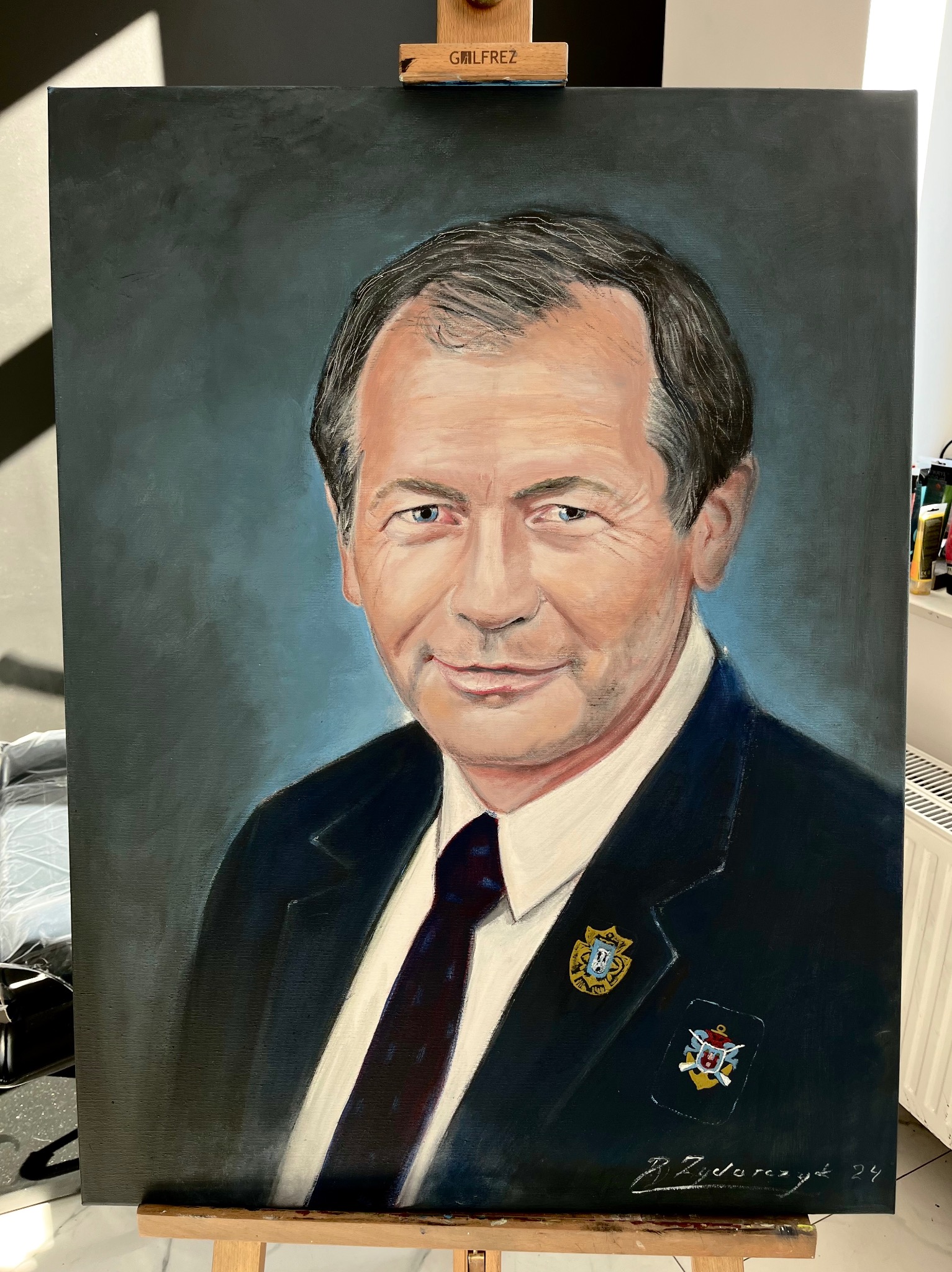 Portret olejny mężczyzny w garniturze z przypinkami, na płótnie, sygnowany w prawym dolnym rogu, ustawiony na sztalugach.