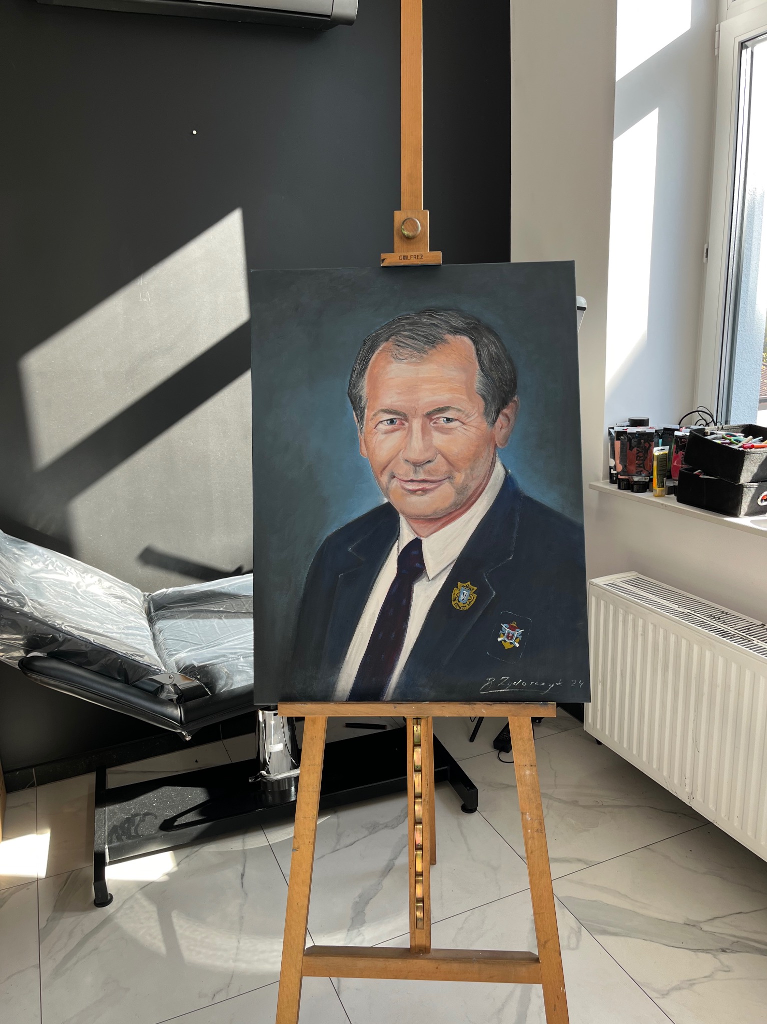 Portret olejny mężczyzny w garniturze, z detalami odznaczeń, ustawiony na drewnianej sztaludze w jasnym pomieszczeniu, obok leżanki kosmetycznej i okna.