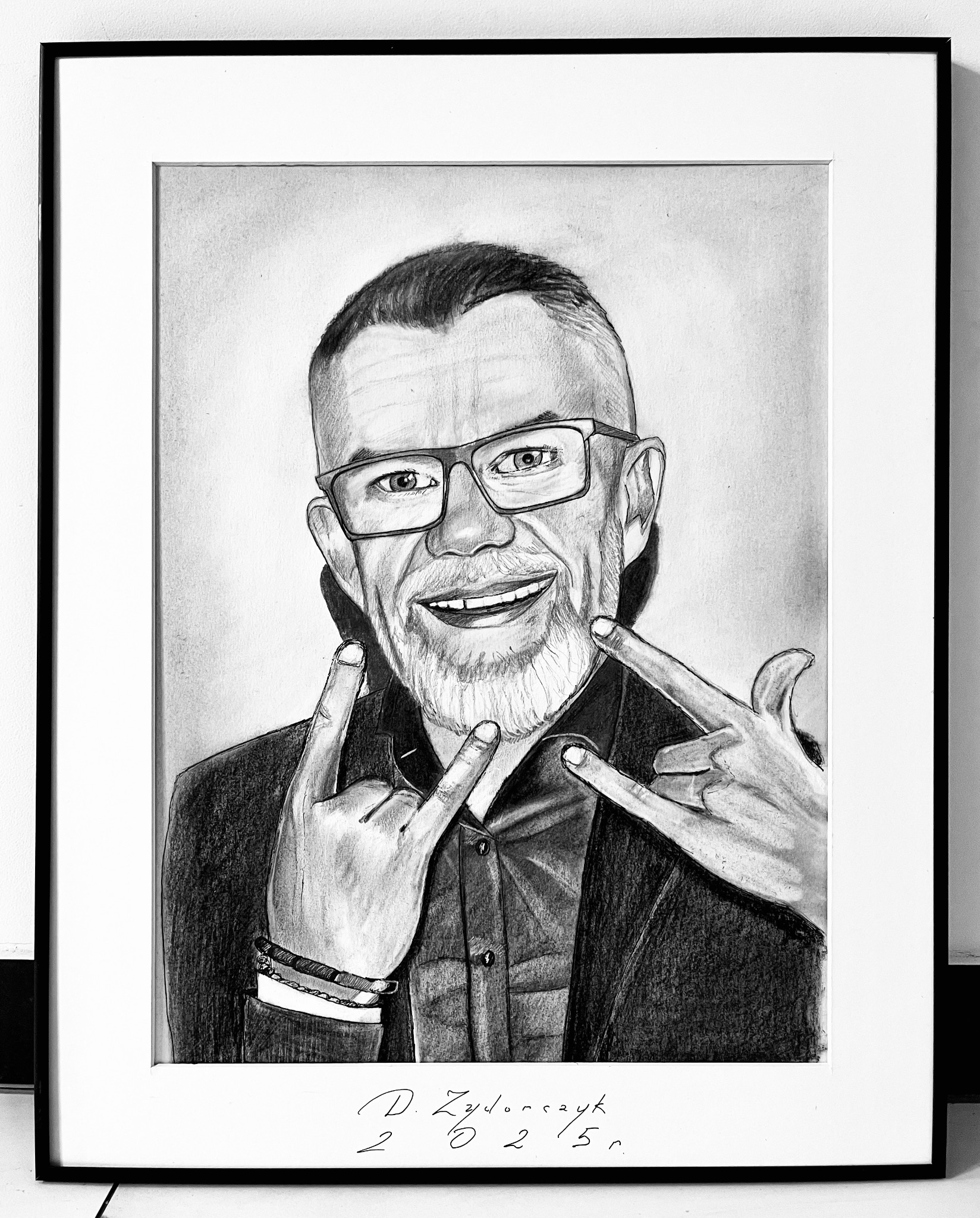 Czarno-biały portret mężczyzny w okularach, wykonany ołówkiem, w czarnej ramce, z uśmiechem i gestem 'rock on' obiema rękami.