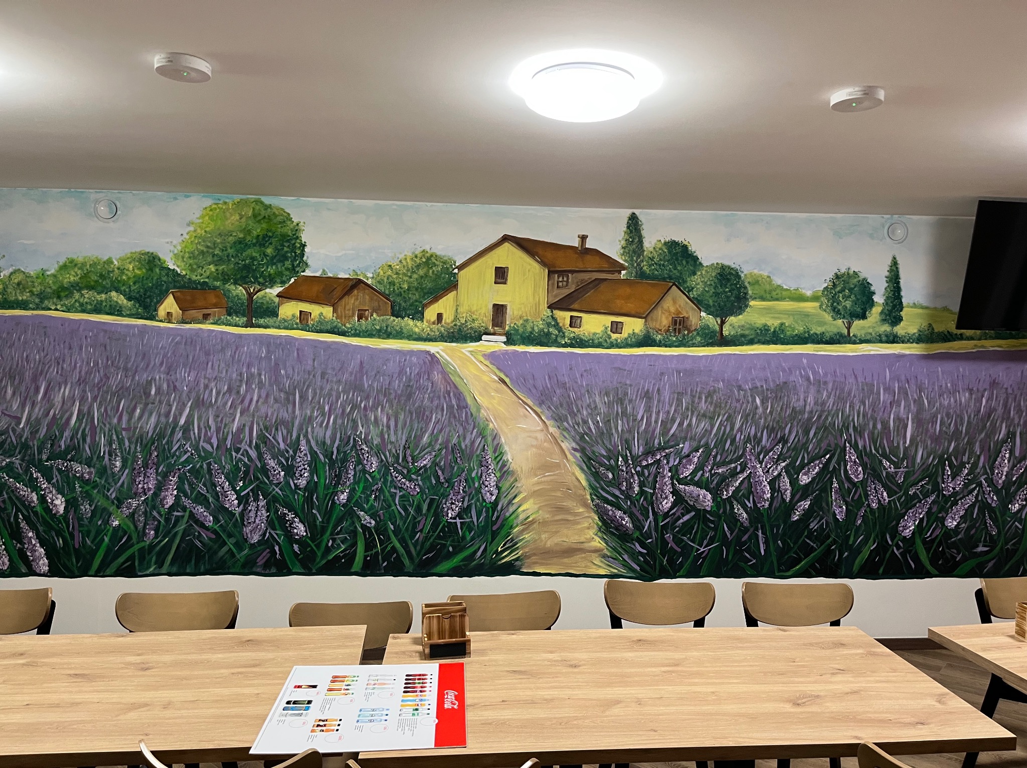 Ścienne malowidło przedstawiające pejzaż z lawendowym polem i domami, widoczne zza stołu z krzesłami w pomieszczeniu. Dekoracja wnętrza z artystycznym akcentem.