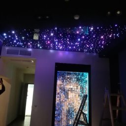 Reghome Sp. z o.o. - Oświetlenie sufitowe imitujące rozgwieżdżone niebo w ciemnym pomieszczeniu, z dekoracyjnym panelem wodnym na ścianie i drabiną opartą o ścianę.