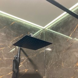 Reghome Sp. z o.o. - Nowoczesna kabina prysznicowa z czarną deszczownicą na tle płytek imitujących marmur z złotymi żyłami i podświetlanym sufitem.