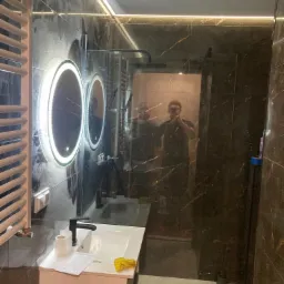 Nowoczesna łazienka z ciemnymi marmurowymi płytkami, podświetlanymi okrągłymi lustrami i grzejnikiem drabinkowym, widoczne odbicie dwóch osób w lustrze kabiny prysznicowej.