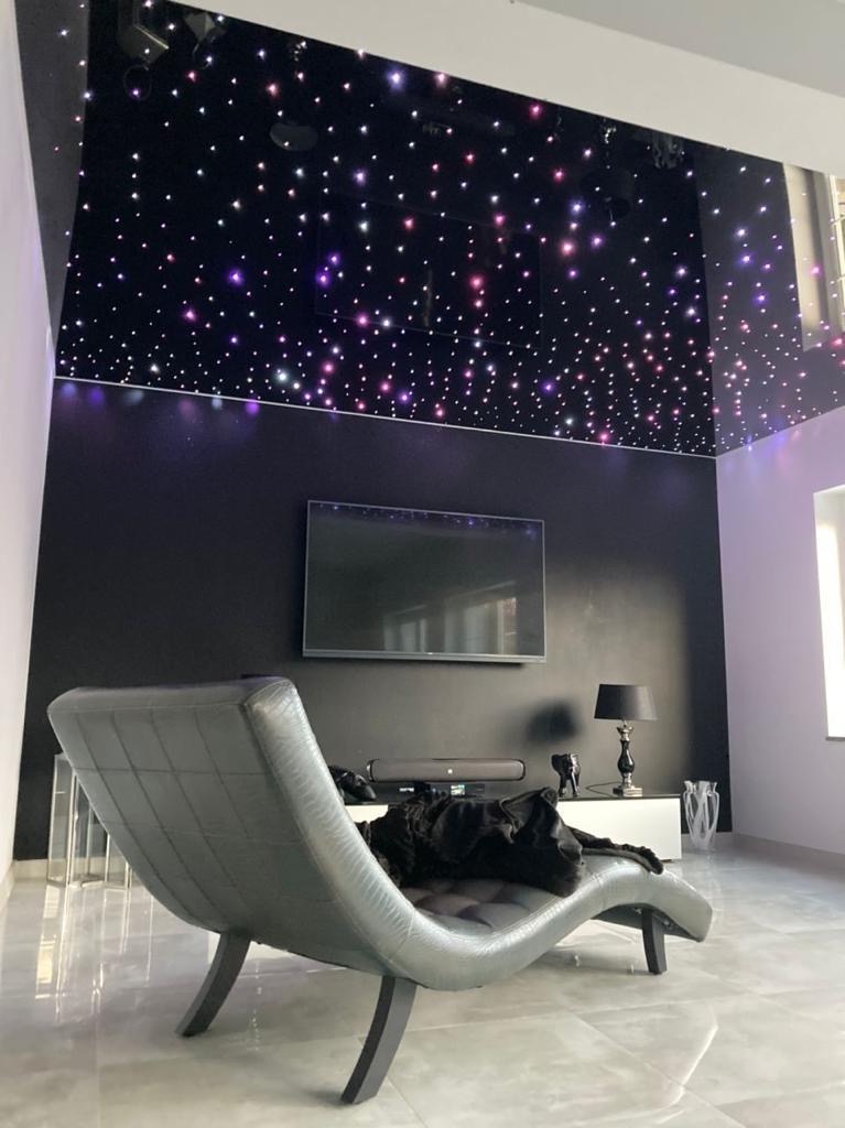 Nowoczesny salon z czarnym sufitem napinanym w gwiazdy, szary fotel relaksacyjny z psem, czarna ściana z telewizorem.