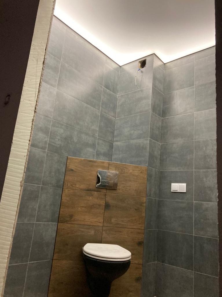 Nowoczesna toaleta podwieszana z szarymi płytkami i drewnopodobną zabudową, widoczne niedokończone wykończenie ściany po lewej stronie.