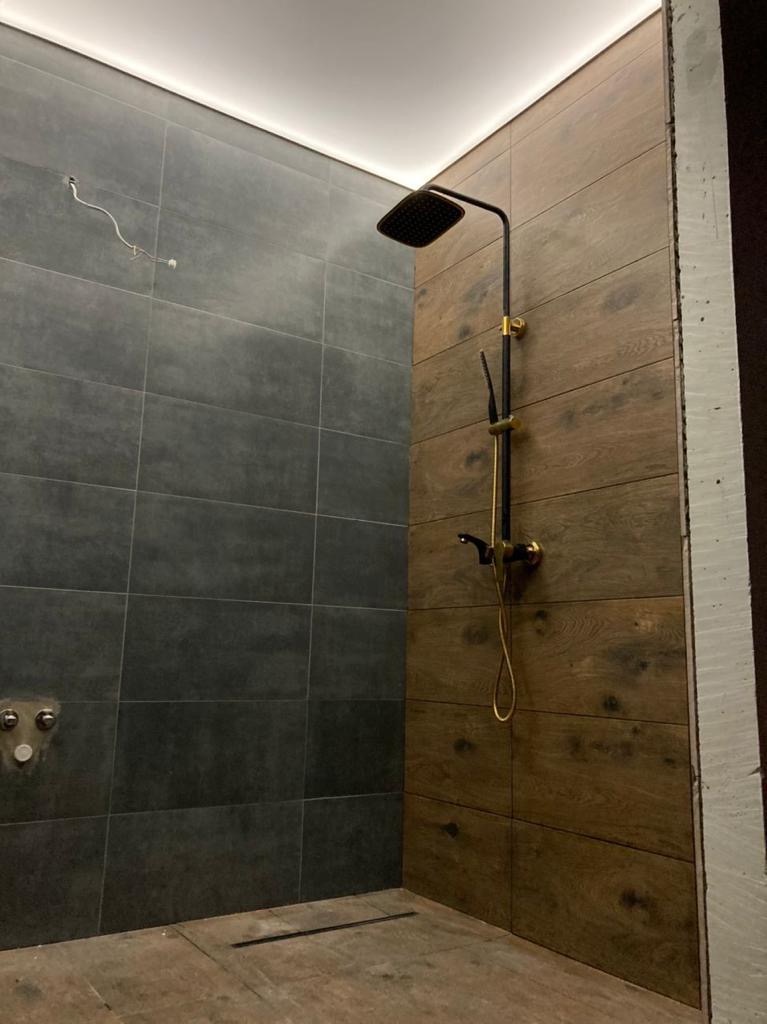 Nowoczesna kabina prysznicowa z czarną armaturą i dwoma rodzajami płytek ściennych: ciemnoszare płytki na jednej ścianie i płytki imitujące drewno na drugiej. Widoczne niedokończone prace...