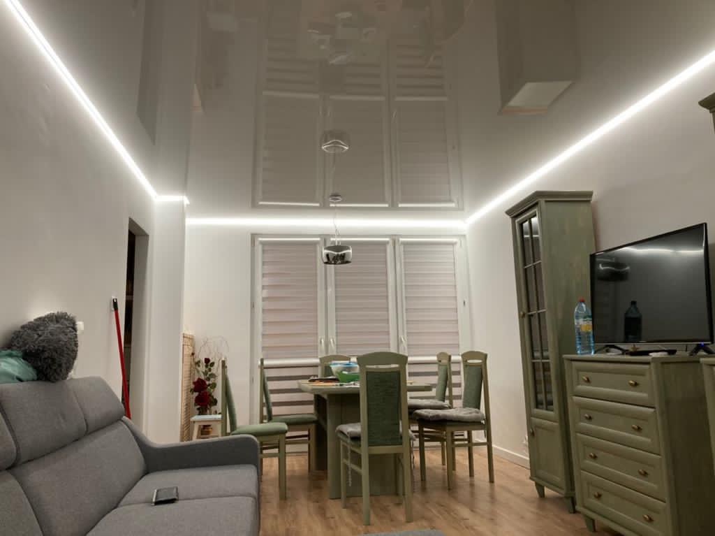 Jasne wnętrze pokoju dziennego z błyszczącym sufitem podwieszanym i listwami LED, widoczny szary narożnik, stół z krzesłami i meble w odcieniach zieleni.