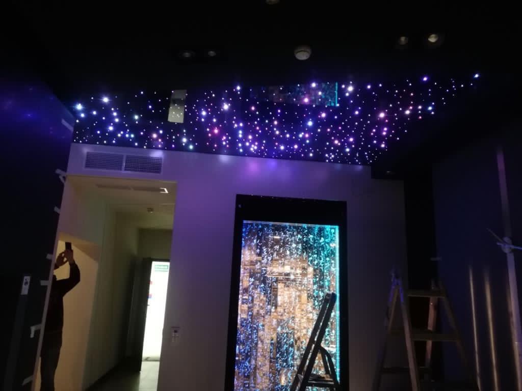 Oświetlenie sufitowe imitujące rozgwieżdżone niebo w ciemnym pomieszczeniu, z dekoracyjnym panelem wodnym na ścianie i drabiną opartą o ścianę.
