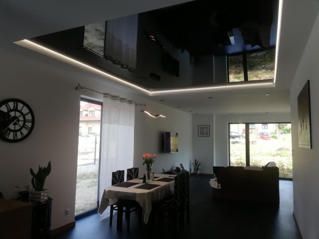 Nowoczesne wnętrze z czarnym, błyszczącym sufitem napinanym i oświetleniem LED, odbijającym widok z okien, stół jadalny z kwiatami, zegar na ścianie.