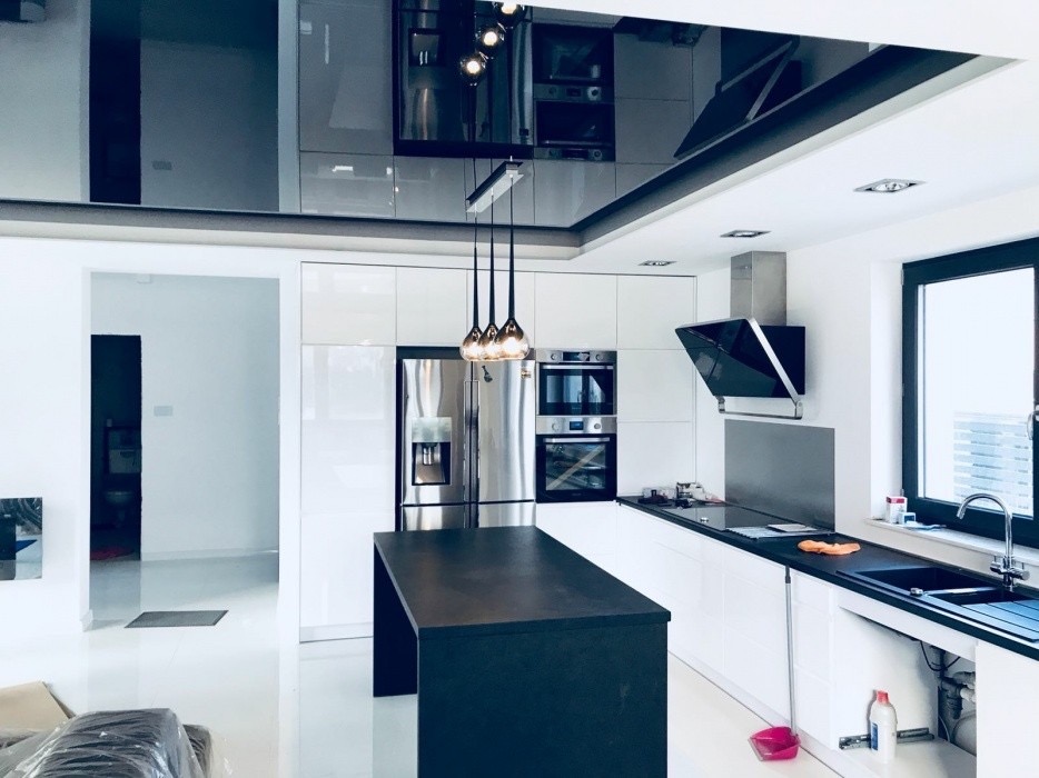 Nowoczesna, minimalistyczna kuchnia z czarnym, lustrzanym sufitem, białymi szafkami, wyspą i sprzętem AGD ze stali nierdzewnej, oświetlona wiszącymi lampami z bursztynowego szkła.
