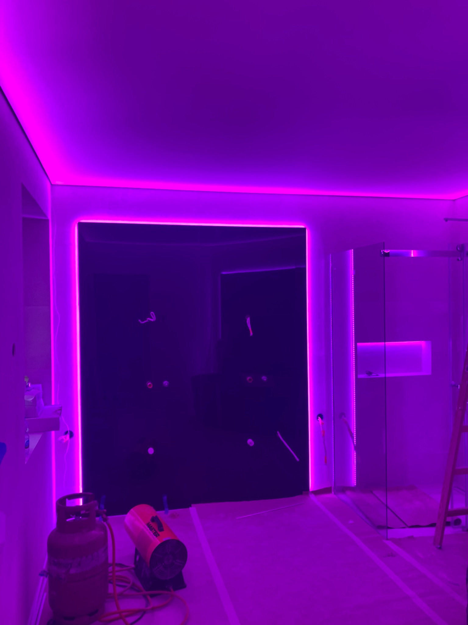 Łazienka w trakcie remontu z podświetleniem LED w kolorze fioletowym, widoczny prysznic ze szklaną kabiną, grzejnik budowlany i butla gazowa.
