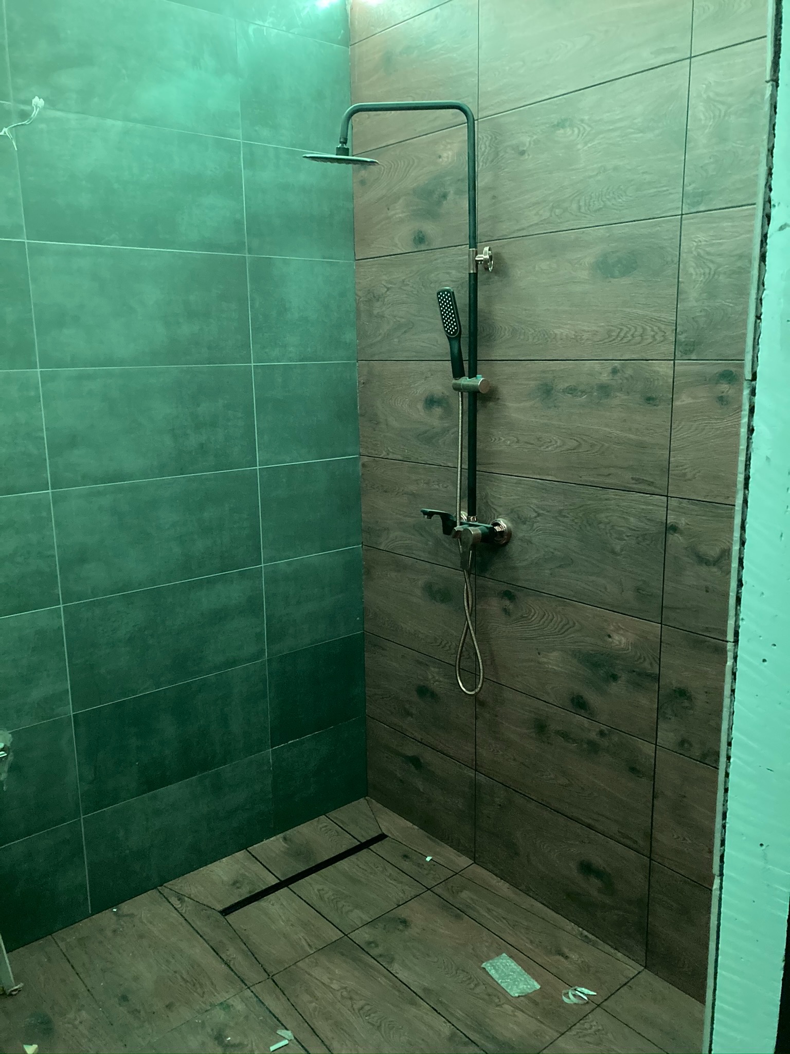 Nowoczesna kabina prysznicowa z ciemnozielonymi i drewnopodobnymi płytkami, czarną armaturą i odpływem liniowym w podłodze, w trakcie wykańczania.