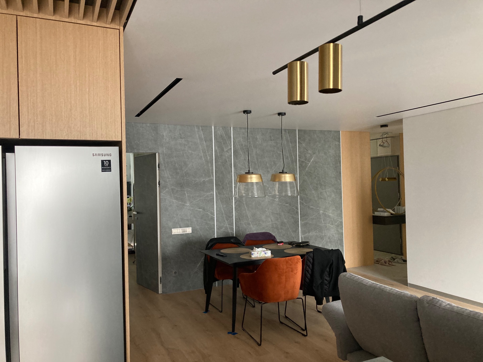 Nowoczesne wnętrze jadalni z szarą ścianą imitującą marmur, drewnianą zabudową kuchenną z lodówką Samsung, czarnym stołem otoczonym krzesłami w odcieniach pomarańczy i purpury oraz złotymi lampami...