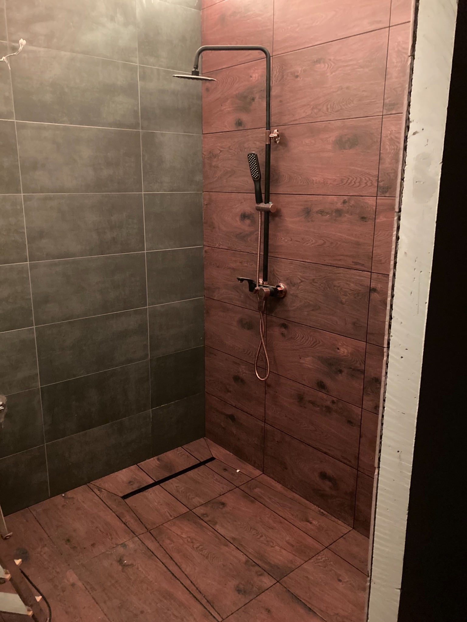 Nowoczesna kabina prysznicowa z brązowymi płytkami imitującymi drewno na ścianach i podłodze, czarny odpływ liniowy, szara ściana obok, czarna armatura prysznicowa z elementami w kolorze miedzi.