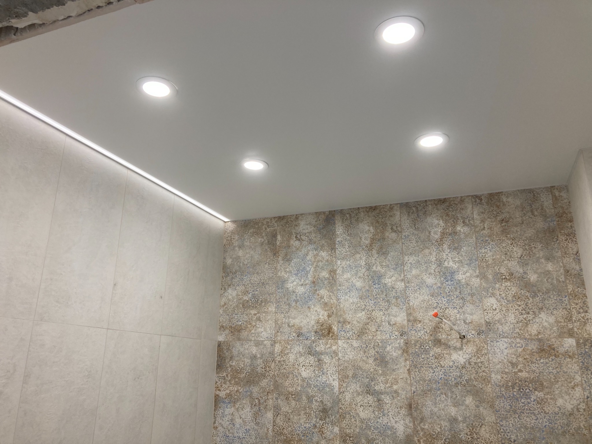 Nowoczesna łazienka po remoncie z białym sufitem podwieszanym z oświetleniem LED, płytki ścienne w odcieniach beżu i błękitu, oraz widoczny fragment instalacji elektrycznej.