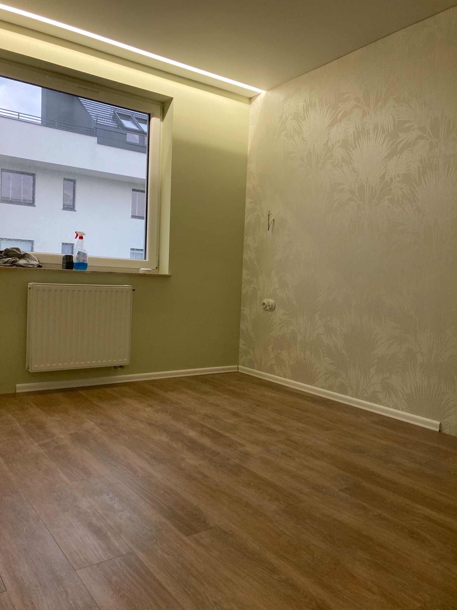 Pokój po remoncie: brązowe panele podłogowe, ściana z tapetą w palmy, grzejnik pod oknem z płynem do szyb i szmatką na parapecie, listwa LED pod sufitem.