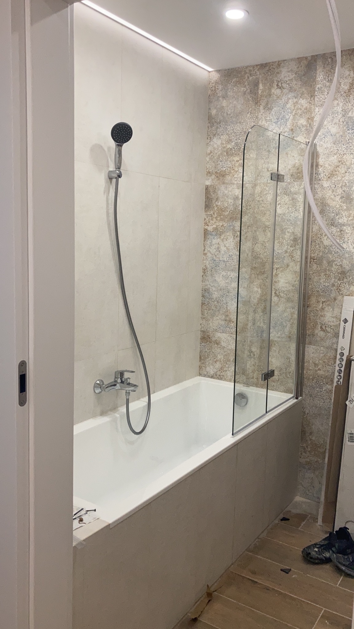 Wykończona łazienka z wanną, szklanym parawanem nawannowym i panelami ściennymi w odcieniach beżu i brązu. Widoczna armatura, w tym bateria i słuchawka prysznicowa. Podwieszany sufit z oświetleniem...