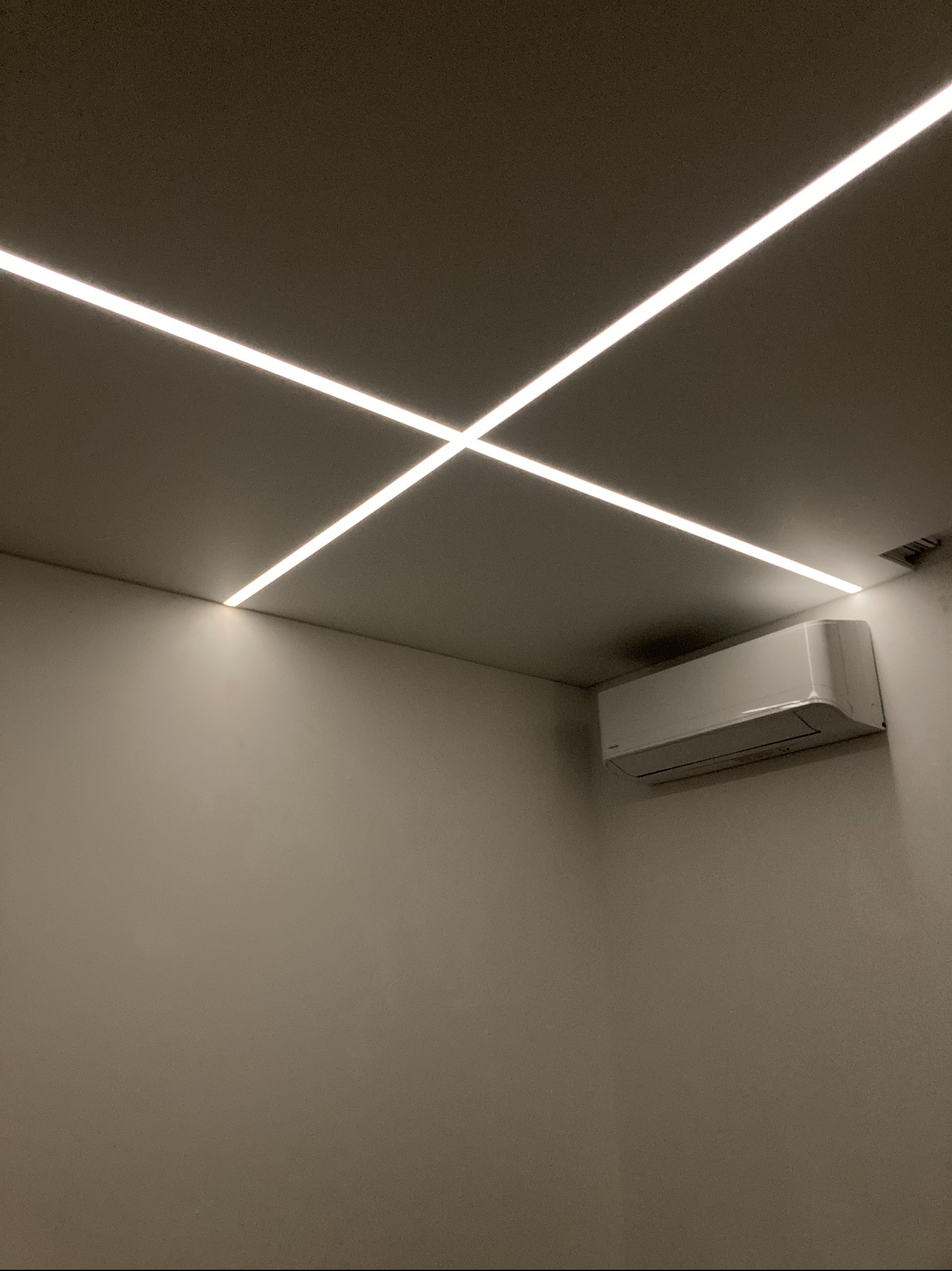 Minimalistyczne wnętrze z krzyżującymi się liniami oświetlenia LED w suficie i klimatyzatorem na ścianie pomalowanej na jednolity kolor.