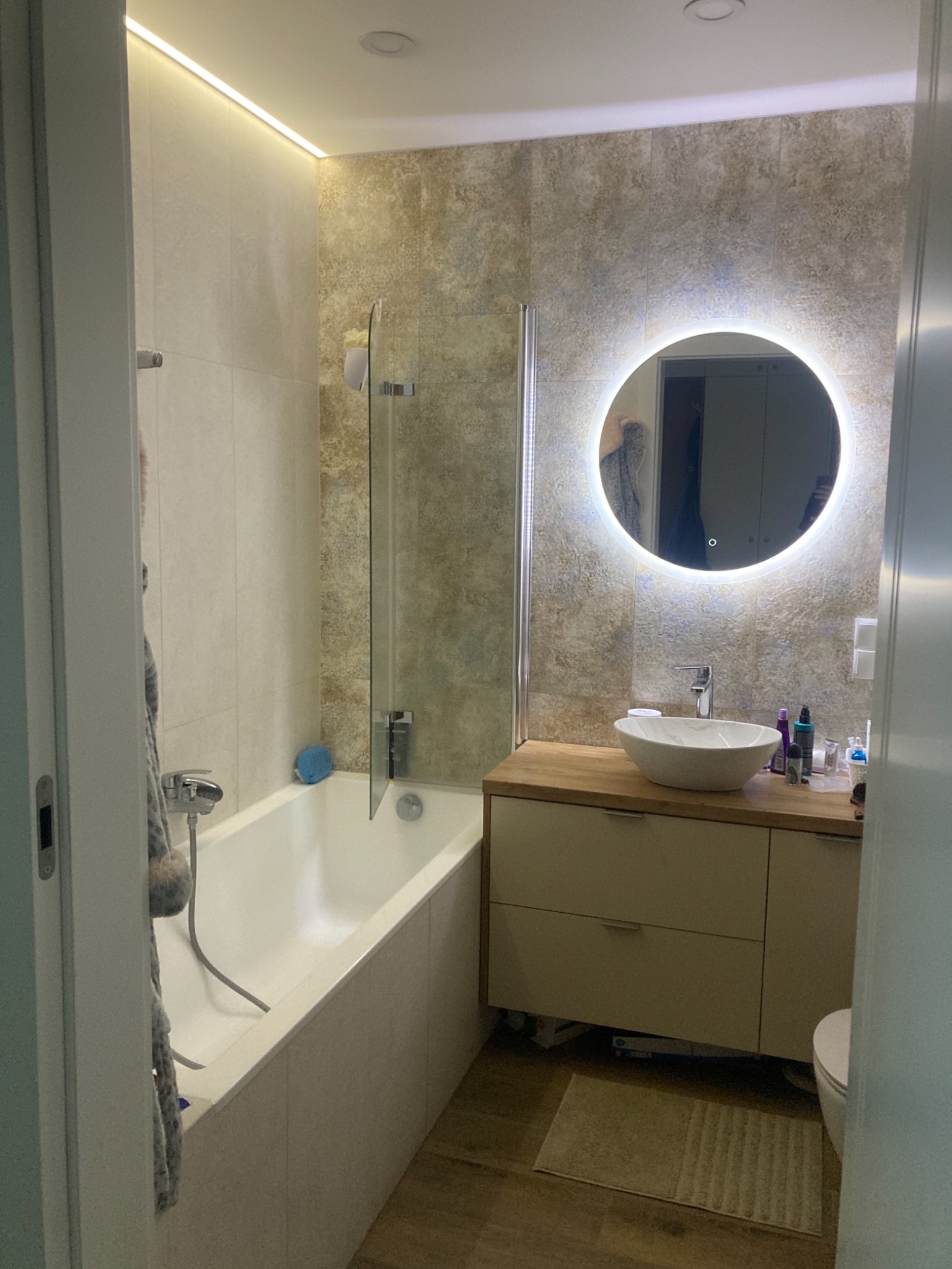 Nowoczesna łazienka z podświetlanym lustrem, umywalką nablatową i wanną z parawanem, wykończona płytkami w odcieniach beżu i brązu, oświetlenie LED podsufitowe.