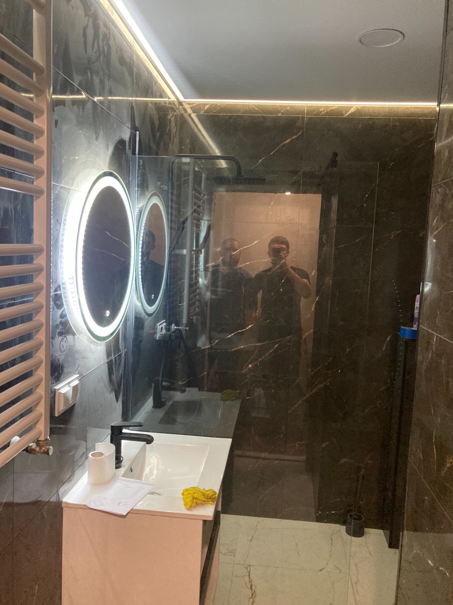 Nowoczesna łazienka z ciemnymi marmurowymi płytkami, podświetlanymi okrągłymi lustrami i grzejnikiem drabinkowym, widoczne odbicie dwóch osób w lustrze kabiny prysznicowej.