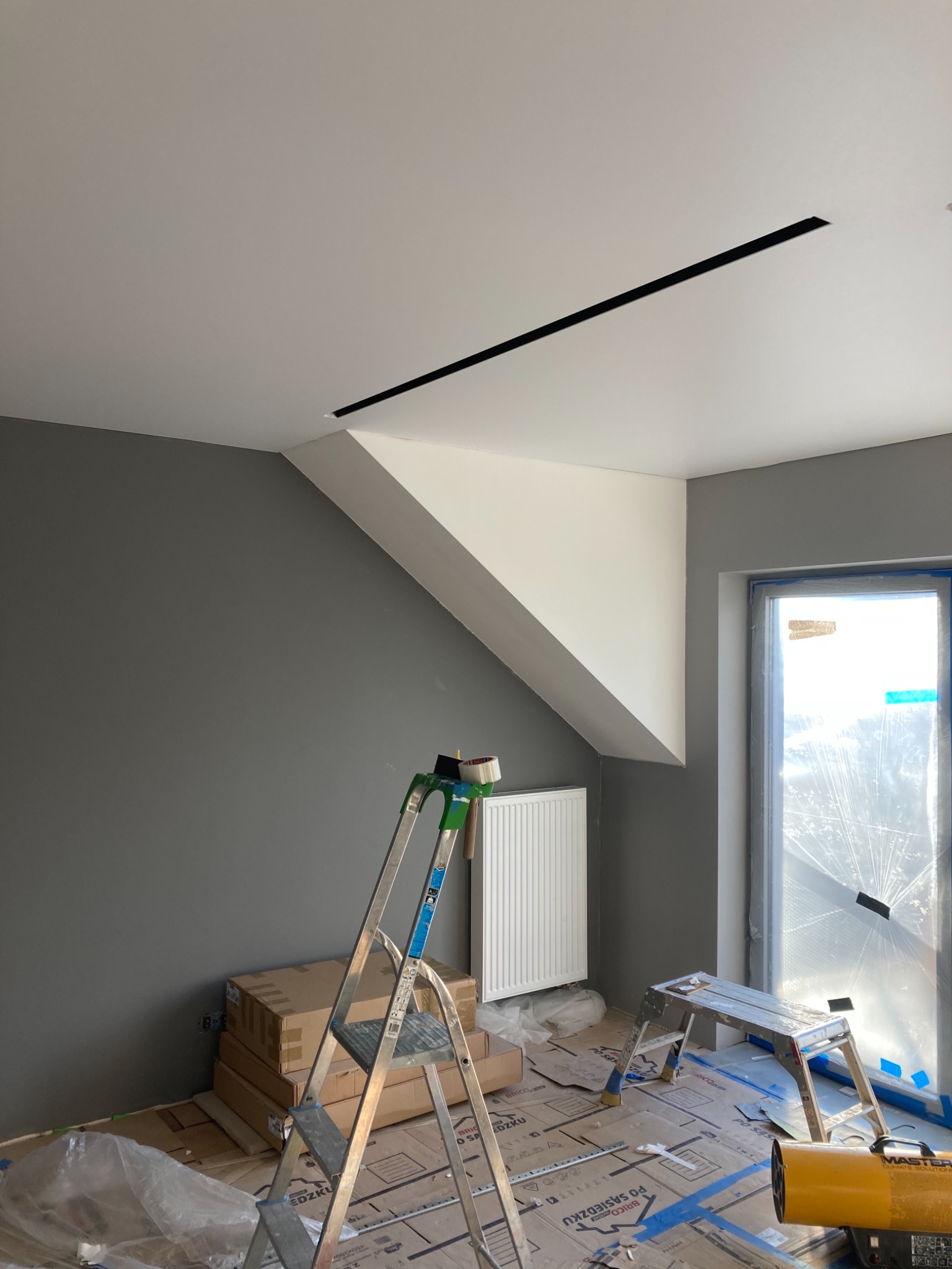 Pokój w trakcie malowania: szara ściana, biały sufit z czarnym paskiem oświetleniowym, zabezpieczone okno folią, kartony na podłodze, drabina z taśmą malarską i grzejnik. Widoczny etap prac...