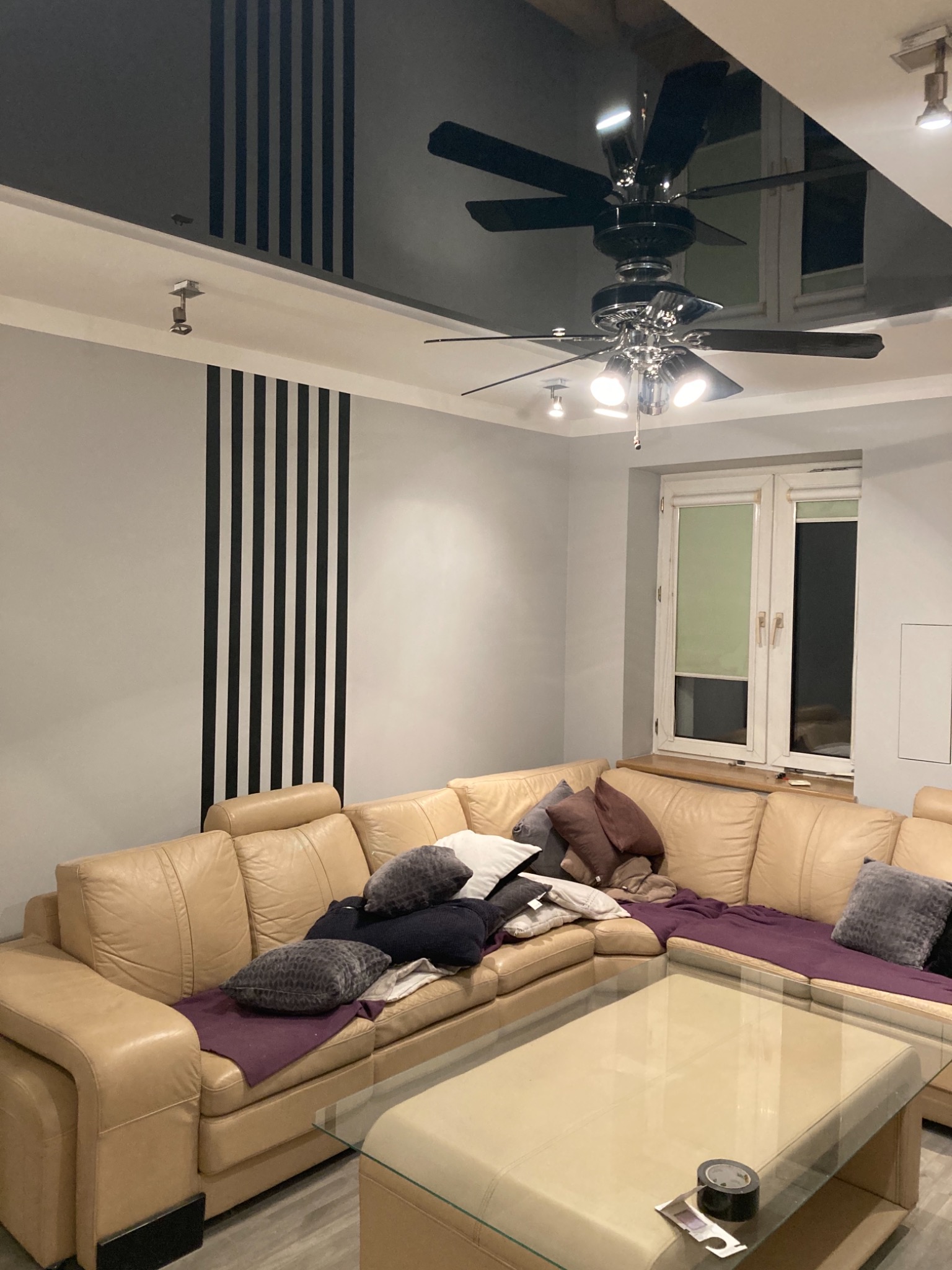 Salon z beżową skórzaną sofą narożną, szklanym stolikiem kawowym z taśmą klejącą na blacie, czarnym sufitem napinanym z wbudowanym oświetleniem i dekoracyjnymi czarnymi pasami na ścianie.