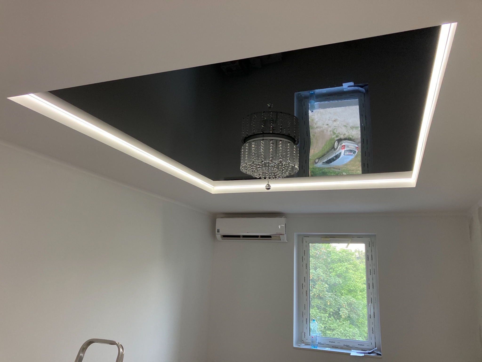 Czarny, lustrzany sufit napinany z oświetleniem LED i kryształowym żyrandolem we Wrocławiu, odbijający widok z okna.