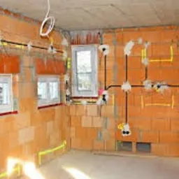 Wnętrze w stanie surowym z widoczną instalacją elektryczną w peszlach, poprowadzoną po ścianach z cegły, z zaznaczonym przebiegiem przewodów żółtą taśmą. Widoczne puszki elektryczne i wystające...