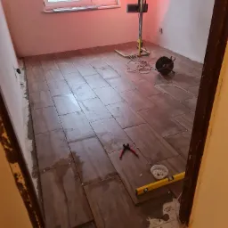 Układanie brązowych płytek podłogowych w pomieszczeniu, widoczne narzędzia glazurnicze: poziomica, szczypce, krzyżyki dystansowe, lampa halogenowa, kabel zasilający i gąbka do czyszczenia fug.