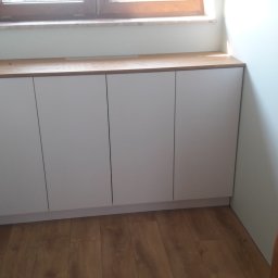 Tomasz Puechowicz P&T - Białe szafki pod oknem z drewnianym blatem, widoczne na tle drewnianej podłogi i ściany z drewnianą belką z włącznikiem światła.