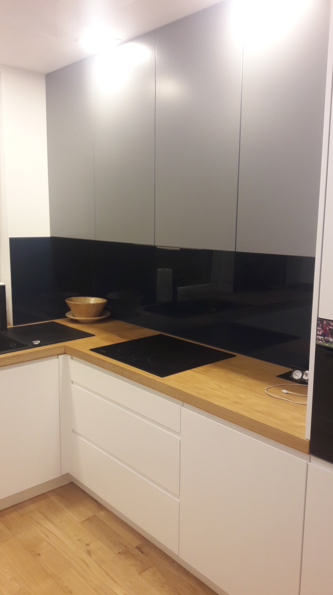 Nowoczesna kuchnia z białymi frontami szafek dolnych i szarymi górnymi, drewniany blat, czarny panel szklany nad blatem, płyta indukcyjna i drewniana misa na blacie.