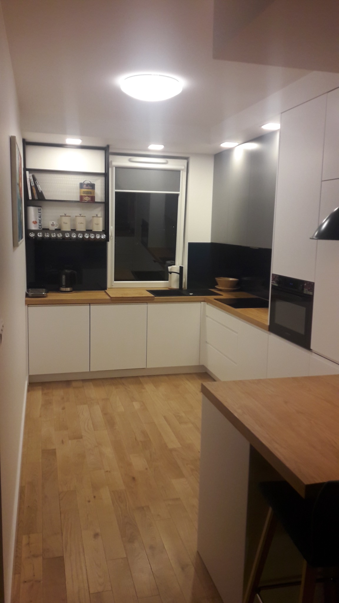 Nowoczesna kuchnia z białymi szafkami, drewnianym blatem i czarnym panelem ściennym, oświetlona światłem sufitowym i punktowym, z widoczną wyspą kuchenną i drewnianą podłogą.