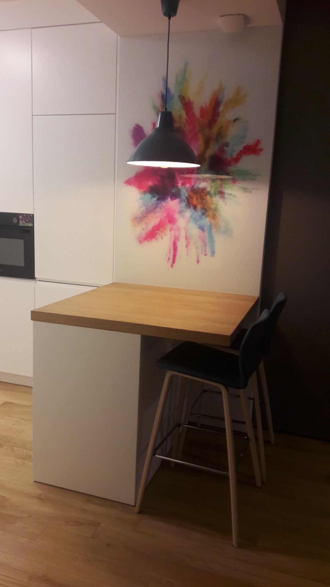 Kompaktowa kuchnia z blatem drewnianym, oświetlona wiszącą lampą, z krzesłami barowymi i dekoracyjnym motywem kolorowych eksplozji na ścianie.
