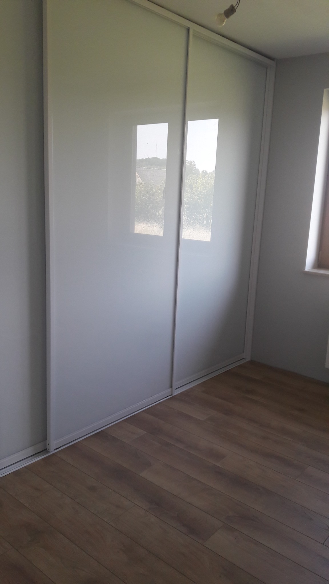 Biała szafa przesuwna z lustrzanymi drzwiami, w której odbija się widok z okna, na tle brązowej podłogi z paneli.