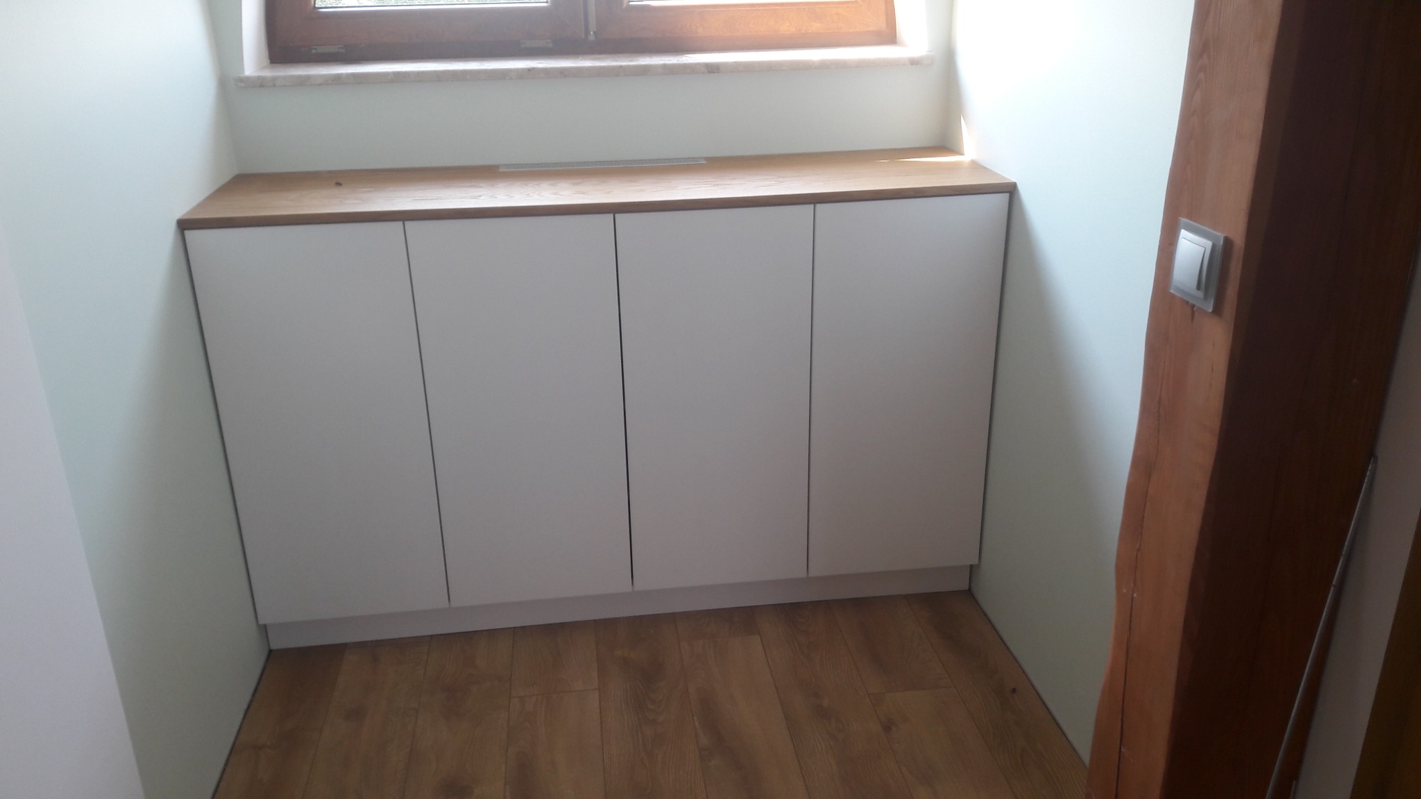 Białe szafki pod oknem z drewnianym blatem, widoczne na tle drewnianej podłogi i ściany z drewnianą belką z włącznikiem światła.