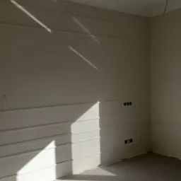 Światło słoneczne wpadające do pokoju z dekoracyjną sztukaterią na ścianach, widoczne gniazdka elektryczne i wystające kable ze ściany.