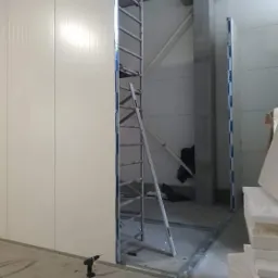 Montaż białych płyt warstwowych PIR w pomieszczeniu przemysłowym, widoczny ruszt metalowy, drabina i wkrętarka na podłodze.