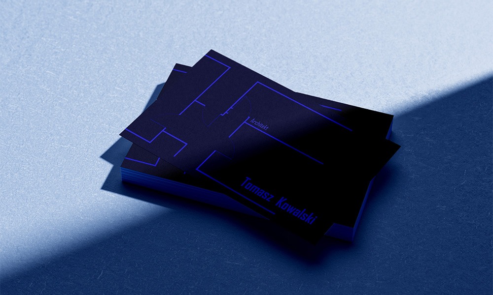 Stos wizytówek architekta w ciemnych odcieniach niebieskiego z minimalistycznym planem budynku i danymi kontaktowymi.