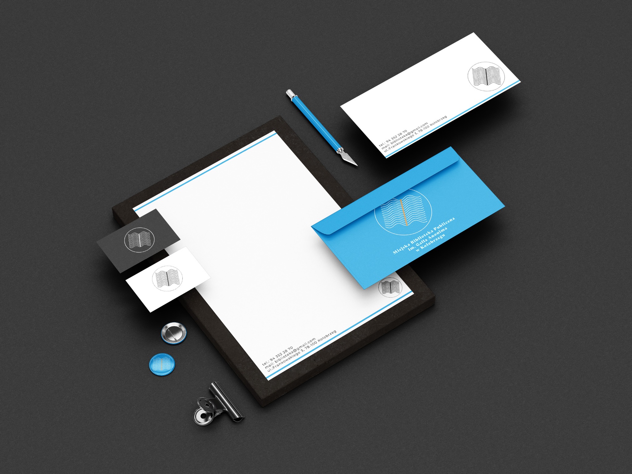 Komplet materiałów firmowych: wizytówki, papier firmowy, koperty z logo Miejskiej Biblioteki Publicznej w Kołobrzegu, ułożone na ciemnym tle z niebieskim nożykiem introligatorskim i metalowymi...