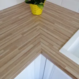 Blat kuchenny z drewna klejonego, ułożonego w jodełkę, w rogu kuchni. Żółte kwiaty w plecionym koszyczku na blacie. Biały zlew ceramiczny w prawym górnym rogu. Białe szafki kuchenne poniżej blatu.