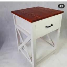 Biały stolik nocny z szufladą i blatem z ciemnego drewna, widoczne charakterystyczne nogi w kształcie litery X, nowoczesny design.