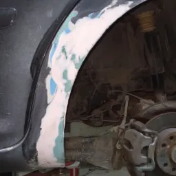 Błotnik samochodu w trakcie naprawy blacharskiej, z nałożoną szpachlą w kolorach białym, zielonym i niebieskim, widoczny element zawieszenia i tarcza hamulcowa, całość na podnośniku samochodowym.