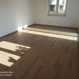 Jasne, nowo wyłożone płytki podłogowe imitujące drewno w pustym pokoju z białymi ścianami i oknem; cień drzewa na podłodze.