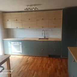 Nowoczesna kuchnia w odcieniach zieleni i drewna w Brzozowie, z białymi płytkami na ścianie i drewnianą podłogą, skórzane uchwyty szafek.