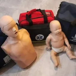 Fantomy osoby dorosłej i dziecka do nauki resuscytacji krążeniowo-oddechowej, torby transportowe i apteczka pierwszej pomocy na szarym tle podłogi.