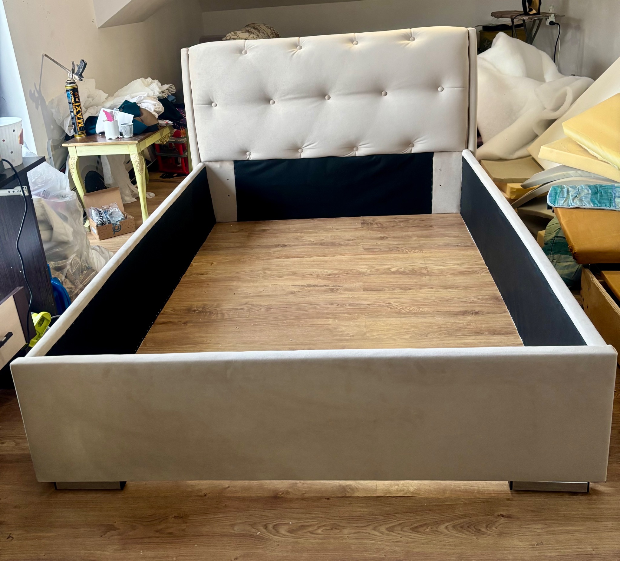 Rama łóżka z tapicerowanym wezgłowiem w odcieniach beżu, widoczne drewniane panele podłogowe i elementy wyposażenia warsztatu w tle.