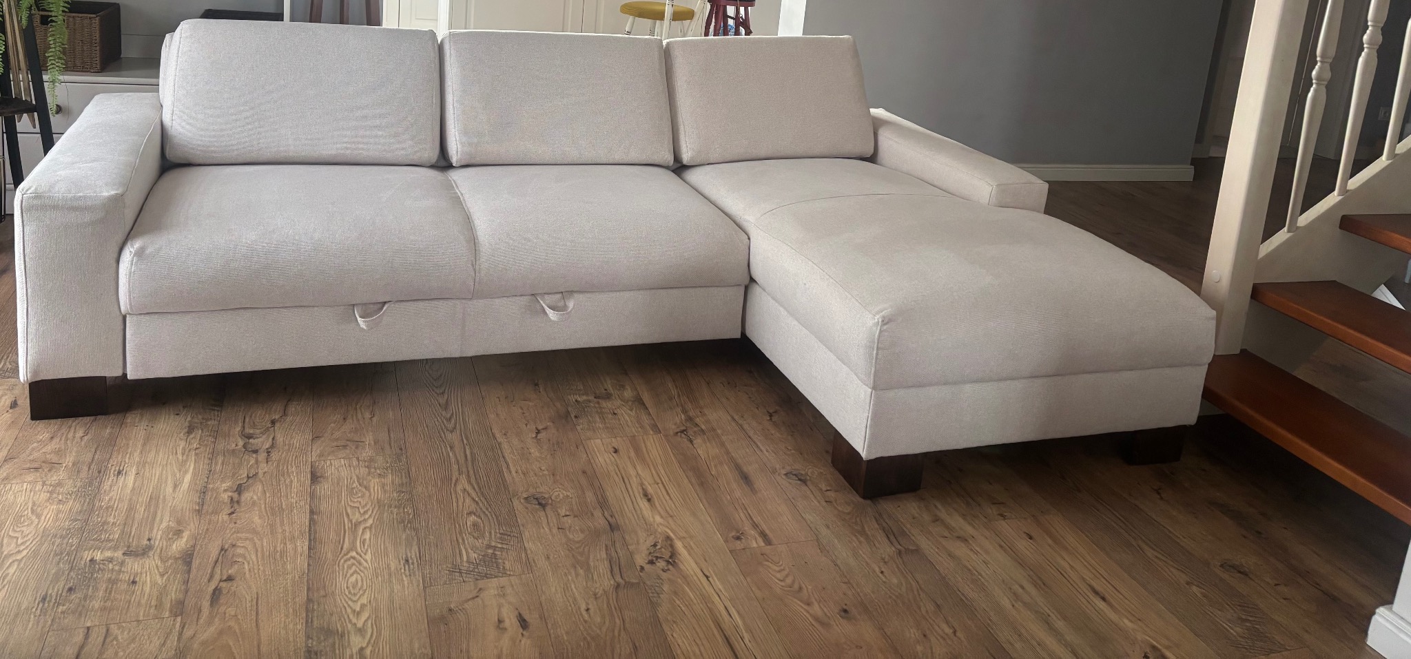 Jasnoszara sofa narożna z funkcją spania i schowkiem, widoczne uchwyty schowka, na drewnianej podłodze, obok schodów.