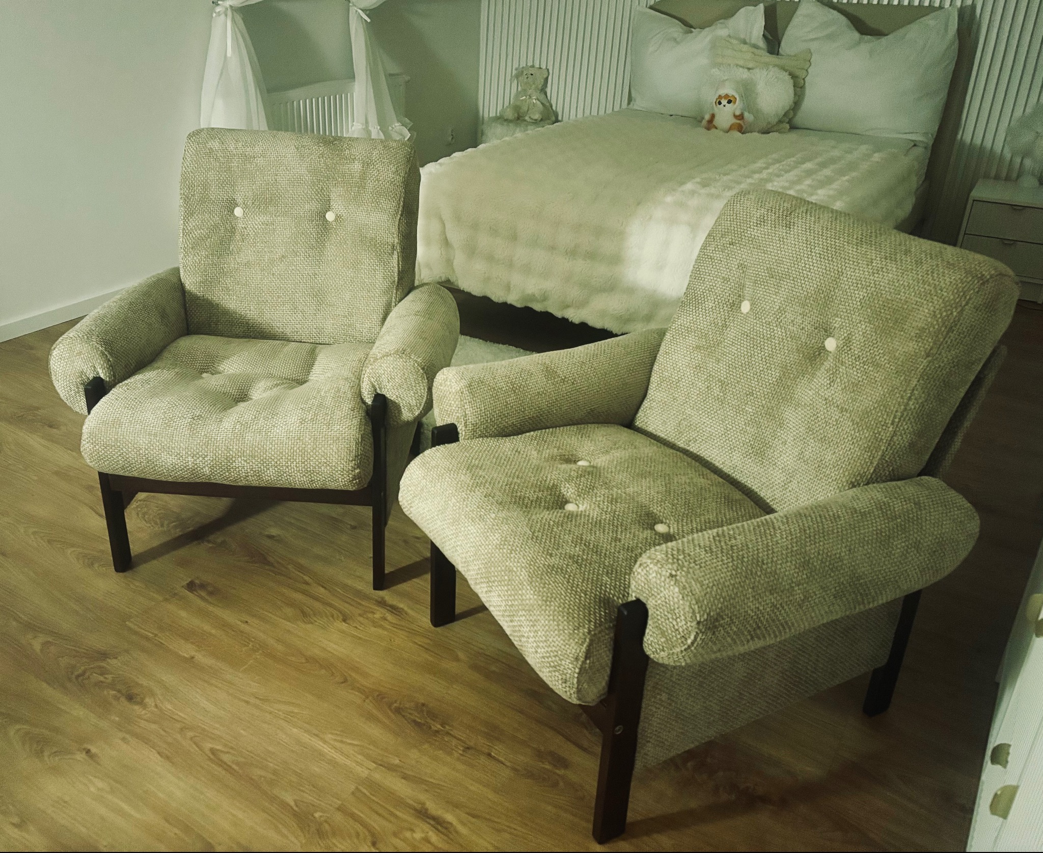 Dwa tapicerowane fotele w stylu retro z brązowymi drewnianymi nogami, ustawione na panelach podłogowych, w tle łóżko z pluszowym kocem i dekoracyjnymi poduszkami.